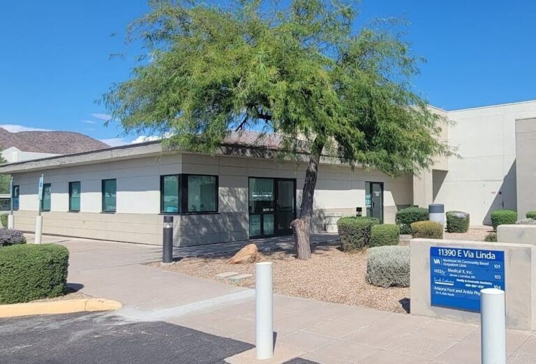 Office building with a large tree leaning over the entrance and a blue directory sign showing the address 11390 E Via Linda